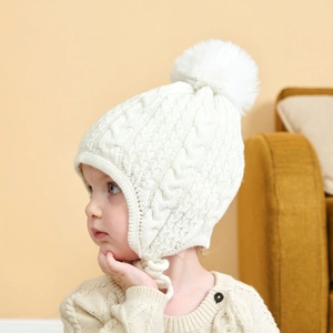 Detail shot of a white knit baby hat featuring a large pompom and soft ear protection