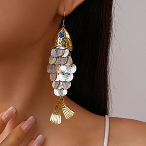 Close-up of golden fish-shaped earring with shell scales and blue eye worn by a model