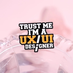 Black enamel badge pin with Trust Me I am a UX/UI Designer quote on glass background