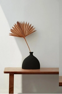 Nahaufnahme der mattschwarzen Keramikvase mit Palmenblatt auf einer Holzbank vor weißer Wand