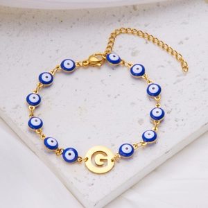 Evil eye bracelet with gold initial G charm on white textured background with silk fabric