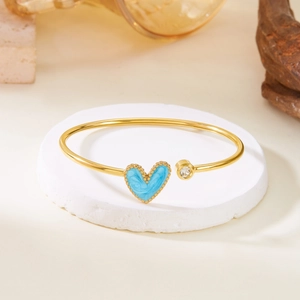 Detail of gold open bangle featuring a blue heart charm with gold beaded edges and zircon
