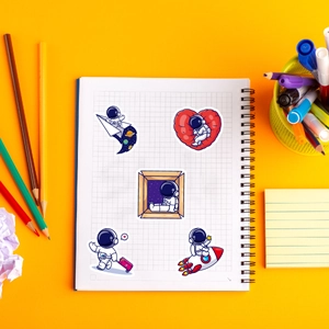 Detail showing five astronaut cartoon stickers on a spiral notebook with art supplies