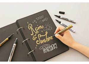 Hand holding a gold Pen-touch paint marker writing on a black notebook with metallic ink