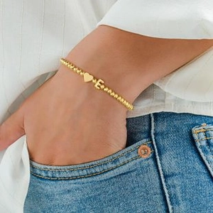 Detail of a gold beaded bracelet with heart and letter E charm worn by a model