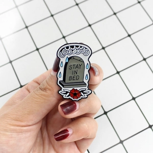 Enamel tombstone badge with flowers and Stay In Bed text held against white background