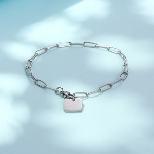 Polished silver paperclip chain bracelet with a square tag charm on a soft blue background