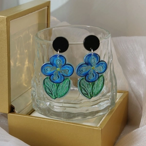 Artistic blue flower earrings with green leaves hanging on a glass cup in a scenic display
