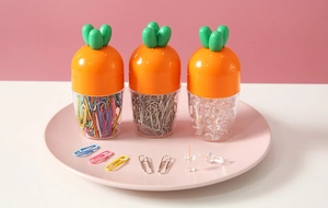 Three-piece carrot-shaped desk organizers with paper clips and push pins on a pink tray