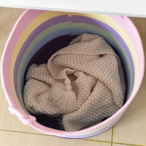Vertical top view of a rainbow cotton rope basket with cream fringed knit blanket inside