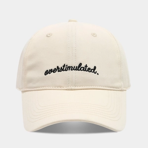 Detail of minimalist beige baseball cap featuring black embroidered overstimulated text