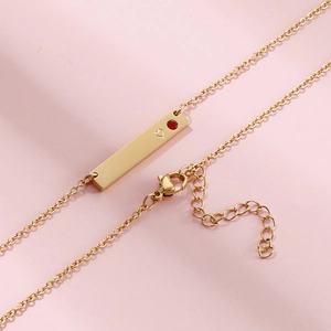 Golden bar pendant necklace with red birthstone and heart detail on pink background