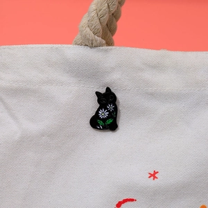 Cute black cat enamel pin with white daisy patterns on a beige canvas bag