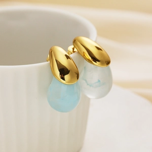 Detail of gold teardrop earrings with light blue resin hanging on a white ceramic cup