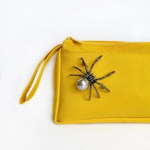 Detail of a yellow canvas pouch featuring a silver metal spider with a large center pearl