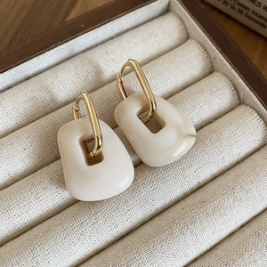 Detailed macro view of square cream acrylic charms on gold earrings in a beige velvet box