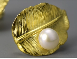 Macro detail of golden feather earrings with a lustrous white pearl on dark background