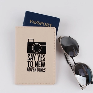 Flat lay of a beige PU leather passport holder with a camera icon and adventure quote