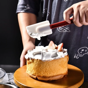 Silicone spatula with red handle spreading cream on cake over a wooden turntable