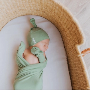 Close-up of sage green newborn swaddle blanket and knotted hat set in a woven bassinet