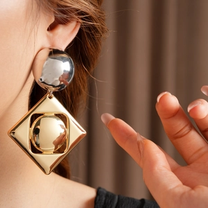 Close-up of geometric earrings featuring a silver sphere and golden hollow square frame