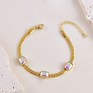 Gold snake chain bracelet with three square iridescent stones on beige marble surface