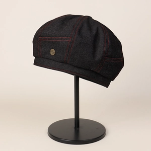 Detail of black denim newsboy cap featuring red contrast stitching and metal badge