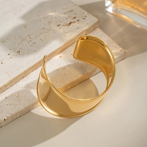 Polished 18K gold-plated wide wave bangle resting on premium textured stone base