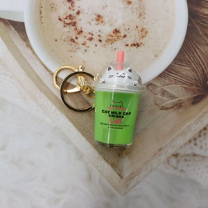 Detail of green cat milk tea keychain with pink straw near a coffee cup on wooden tray
