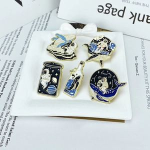 Five creative astronaut and whale enamel pins displayed on a white tray over a magazine