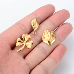 Three gold stainless steel DIY pendants featuring clover, leaf, and shell designs in hand