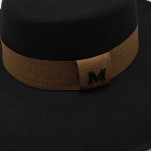 Detail of the brown woven band with a black metal M logo on a black wool felt fedora hat