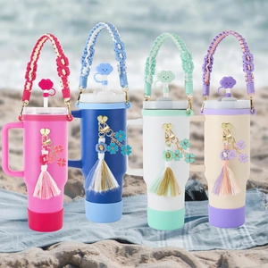 Row of colorful tumblers with woven daisy handles and tassels on a sunny beach