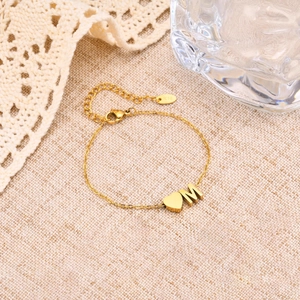 Detail of gold chain bracelet featuring a polished heart and letter M pendant on beige linen