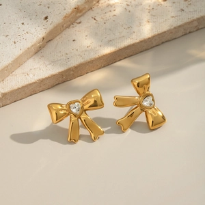 Close-up of golden bow stud earrings with heart-shaped zircon on a beige stone