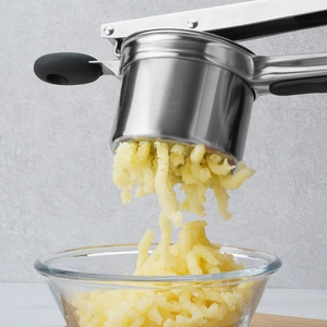 Stainless steel potato masher with black silicone handle pressing potatoes into a glass bowl