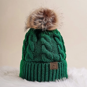 Close-up of dark green cable knit beanie with faux fur pom and leather logo patch