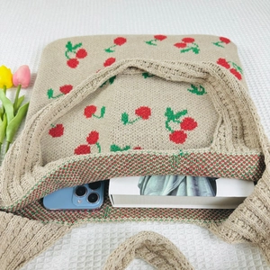 Detail view of beige knit tote bag with cherry pattern containing phone and book