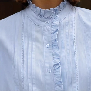 Detail der hellblauen Bluse mit Rüschen-Stehkragen und vertikalen Plissee-Falten