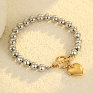 Detalle de una pulsera de cuentas plateadas con colgante de corazón dorado y cierre OT