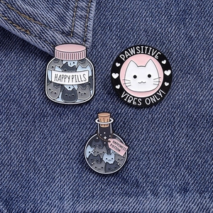 Close-up of three cat enamel pins featuring Happy Pills and Pawsitive Vibes on denim