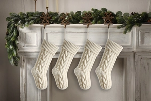 Four white cable-knit Christmas stockings hanging on fireplace with pine branch decor