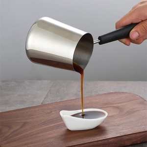Stainless steel milk pan with black handle pouring coffee into a small ceramic dish