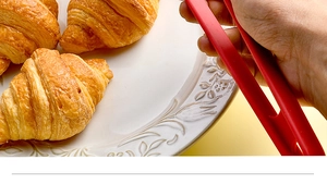 Detail of red silicone food tongs picking up a golden croissant from a white ceramic plate