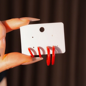 Detailed close-up of glossy red C-shaped hoop earrings on a white display card