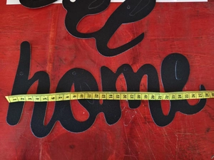 Black metal 'home' wall decor being measured with a yellow tape on a red workbench
