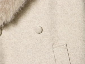 Detail eines beigen Wollmantels mit weicher Textur, rundem Knopf, Tasche und Pelzkragen