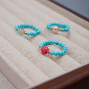 Three handmade turquoise bead rings with pearl and gemstone charms on a beige velvet tray