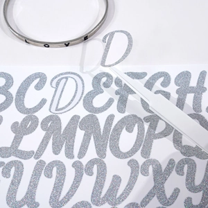 Using tweezers to peel a silver glitter alphabet sticker for DIY graduation cap decoration