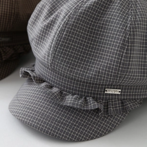 Detail of grey plaid newsboy hat featuring ruffled brim and metallic logo emblem